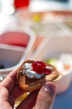 A hand holding a small pancake topped with whipped cream and red jam, with a bokeh background.