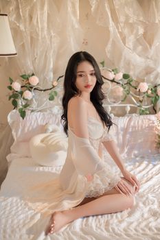 A woman in white lingerie sitting gracefully on a floral-decorated bed in a softly lit room.