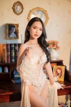Elegant woman in lace lingerie poses indoors with vintage decor in soft lighting.