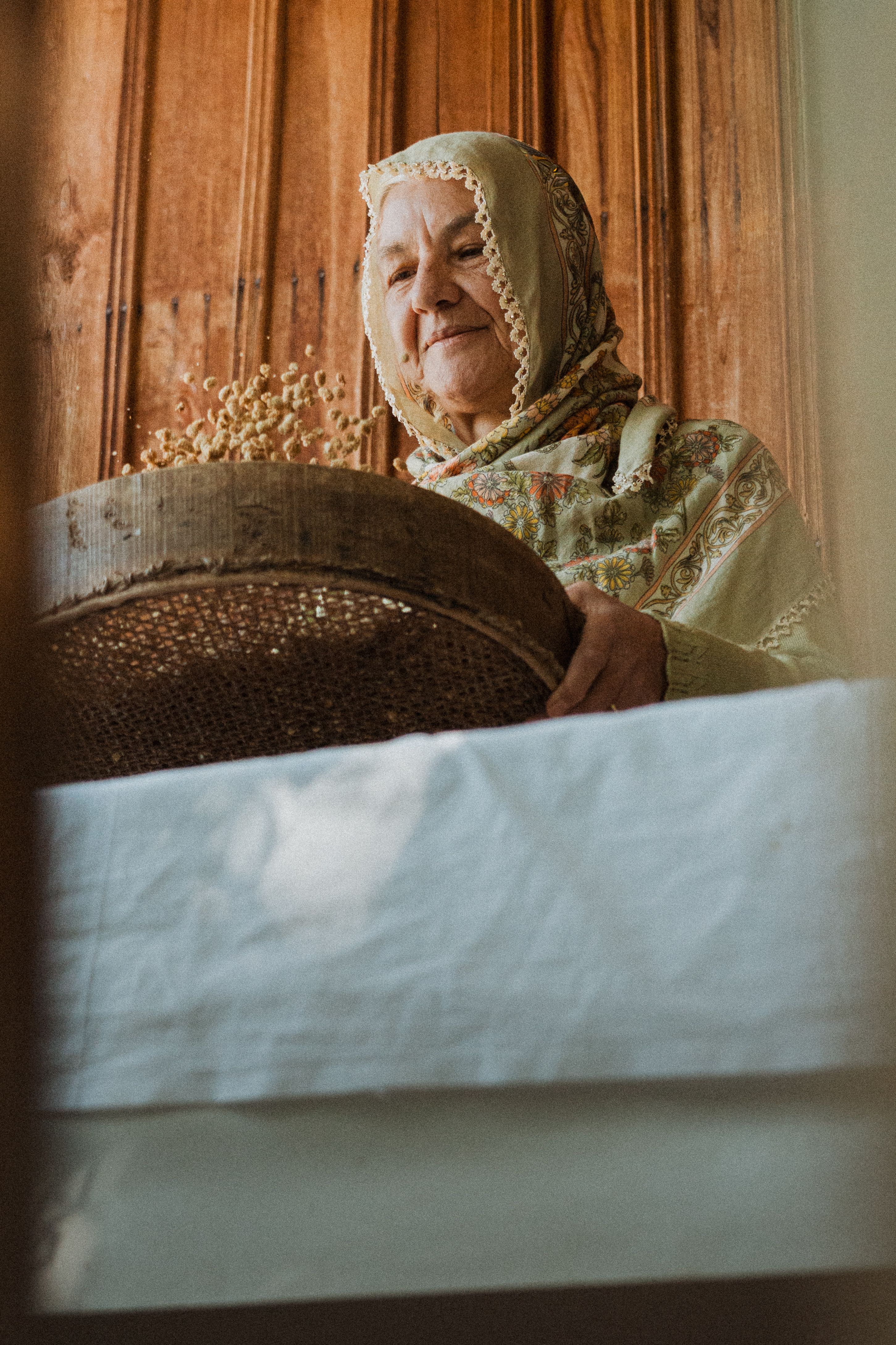 Elderly woman in traditional attire sifting grains inside a rustic home in Erzincan, Türkiye.