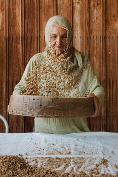 Elderly woman sifting grains with a traditional sieve indoors, rustic setting.