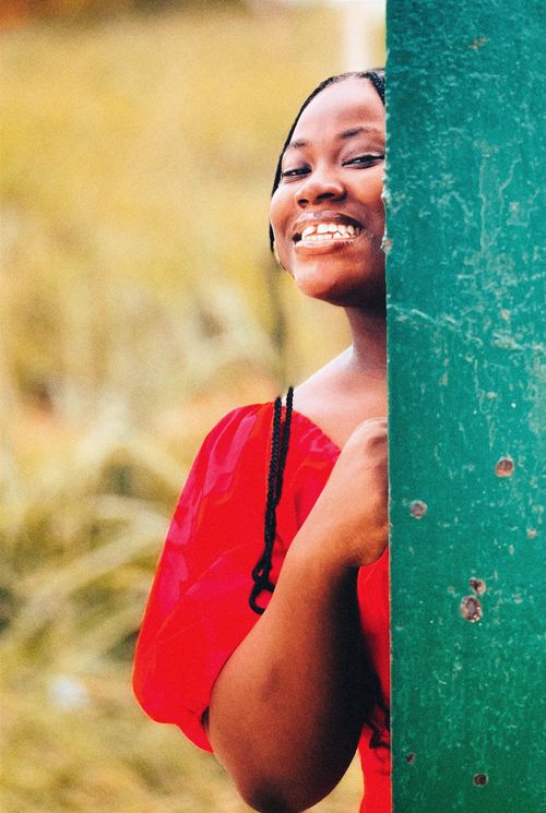 Free Joyful woman in vibrant red dress smiling outdoors, leaning against a post. Stock Photo