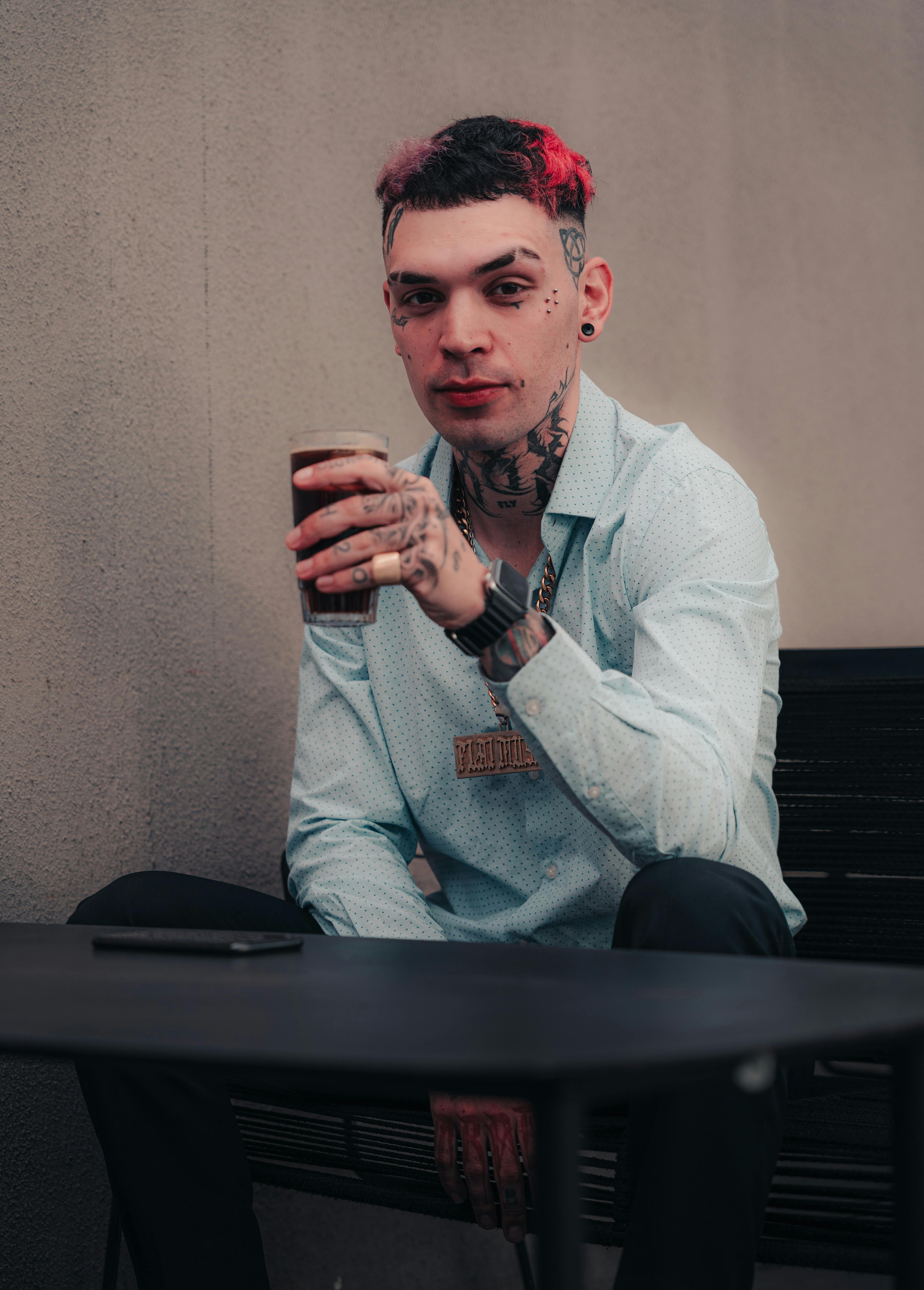 Kostenlos Junger Mann mit Tattoos sitzt im Freien und hält ein Getränk in der Hand, um modernen Stil zu präsentieren. Stock-Foto