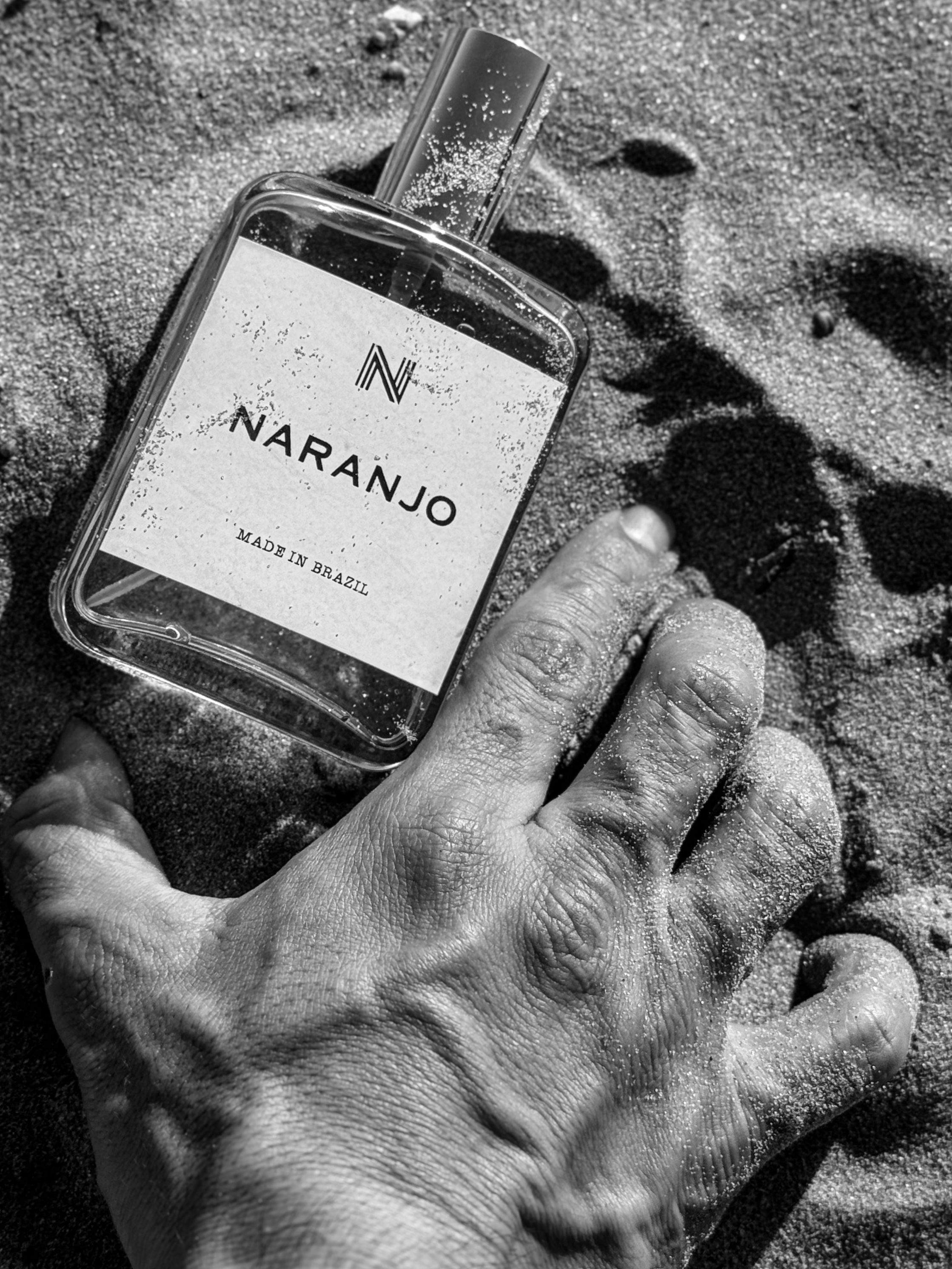 Black and white image of a Naranjo perfume bottle on sand with a male hand.