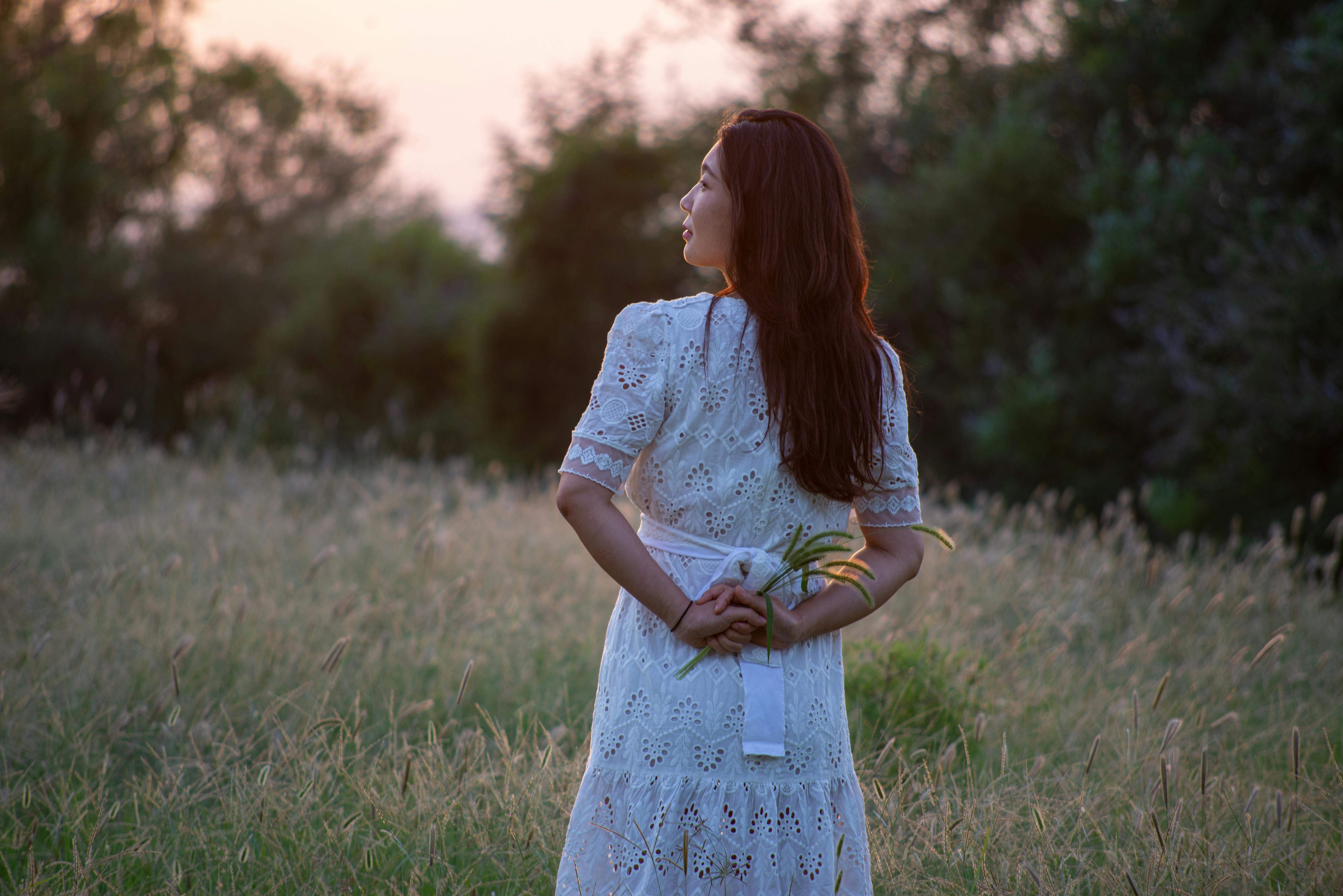 A woman in a white dress holds flowers as she gazes at the sunset in a serene field.
