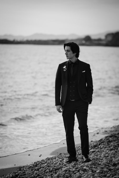 Free A man in a black suit walks along a pebbled beach, captured in a moody black and white tone. Stock Photo