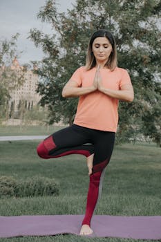 A woman in activewear practices yoga outdoors, performing the tree pose on grass.