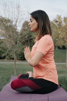 A woman sitting in a yoga pose on a mat in a tranquil outdoor setting, focusing on mindfulness.
