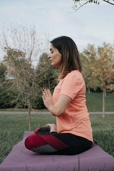 A woman meditating on a yoga mat outside in a park during the day.