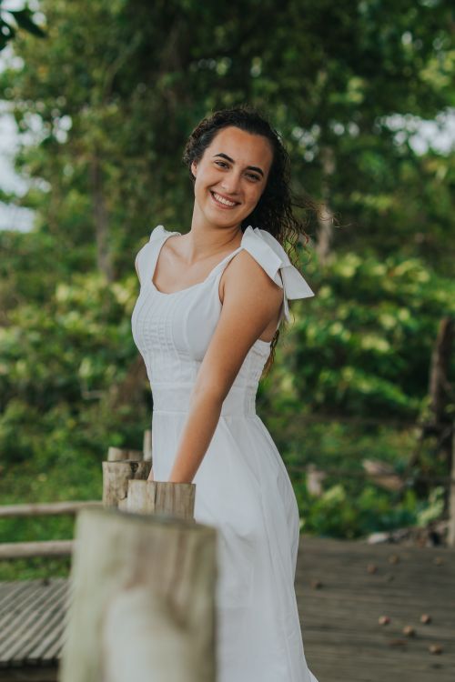 Free A young woman in a white dress smiles while posing outdoors with greenery in the background. Stock Photo