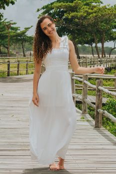 Smiling woman in a flowing white dress walks along a sunny boardwalk surrounded by greenery.