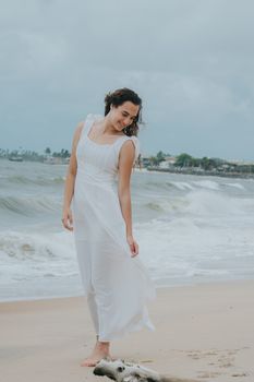 A woman in a flowing white dress stands barefoot on a beach with waves crashing around her, embodying serene elegance.