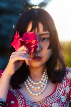 Beautiful woman in traditional attire holding red flowers with a pearl necklace, outdoors at sunset.
