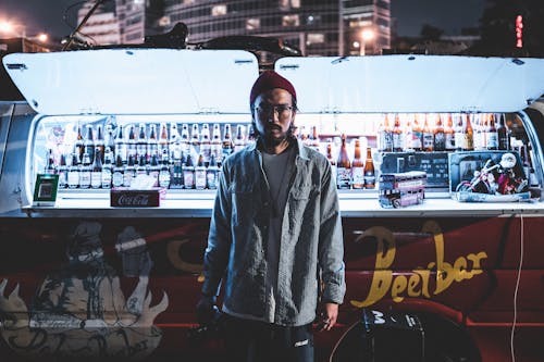 Young Asian male photographer in beanie and glasses wearing casual outfit standing near open street beer bar in night city