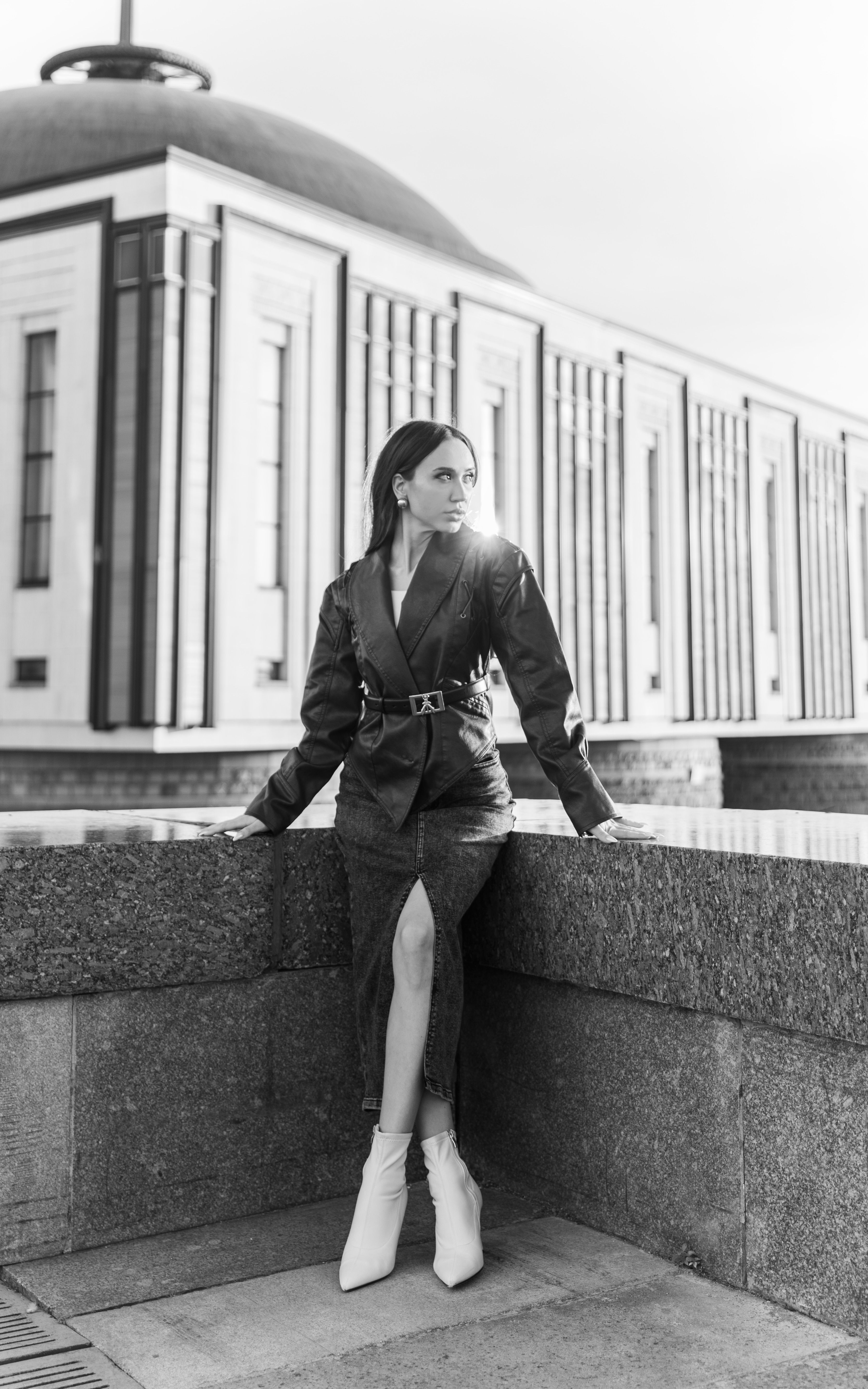 Stylish woman in a blazer and boots poses dramatically in a city backdrop, monochrome effect.