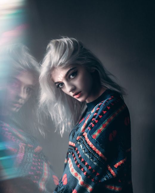 A young woman with striking makeup poses in a reflective portrait showcasing her colorful sweater.