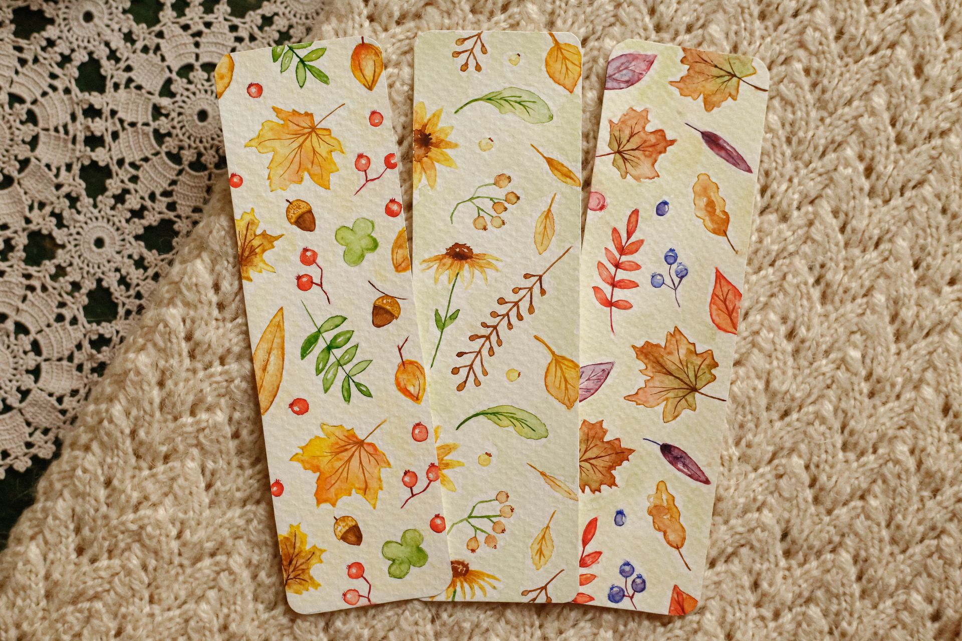 Three autumn-themed bookmarks with watercolor leaves and plants on a cozy knitted surface.