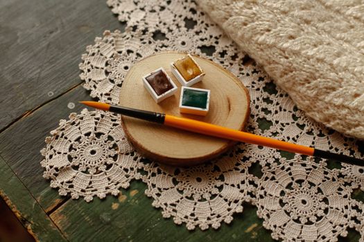 Cozy still life featuring paintbrush, watercolor pans, crochet doilies on wooden surface.