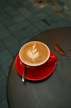 A beautifully crafted latte with heart-shaped art in a red cup on an outdoor table.