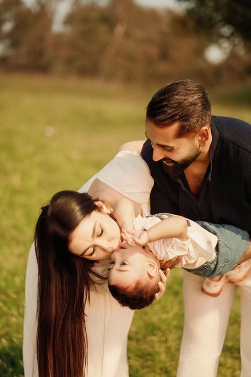 Free A joyful family moment with parents playing and kissing their baby in a sunlit park. Stock Photo