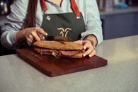 Close-up of a chef slicing a fresh gourmet sandwich on a wooden cutting board indoors.