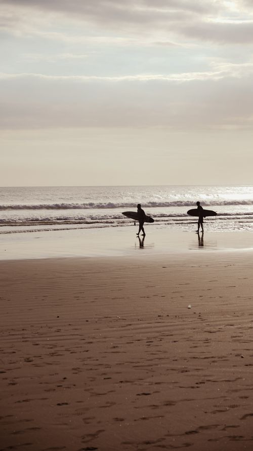 Free Two silhouettes of surfers carrying boards along the beach during sunset, reflecting tranquility. Stock Photo
