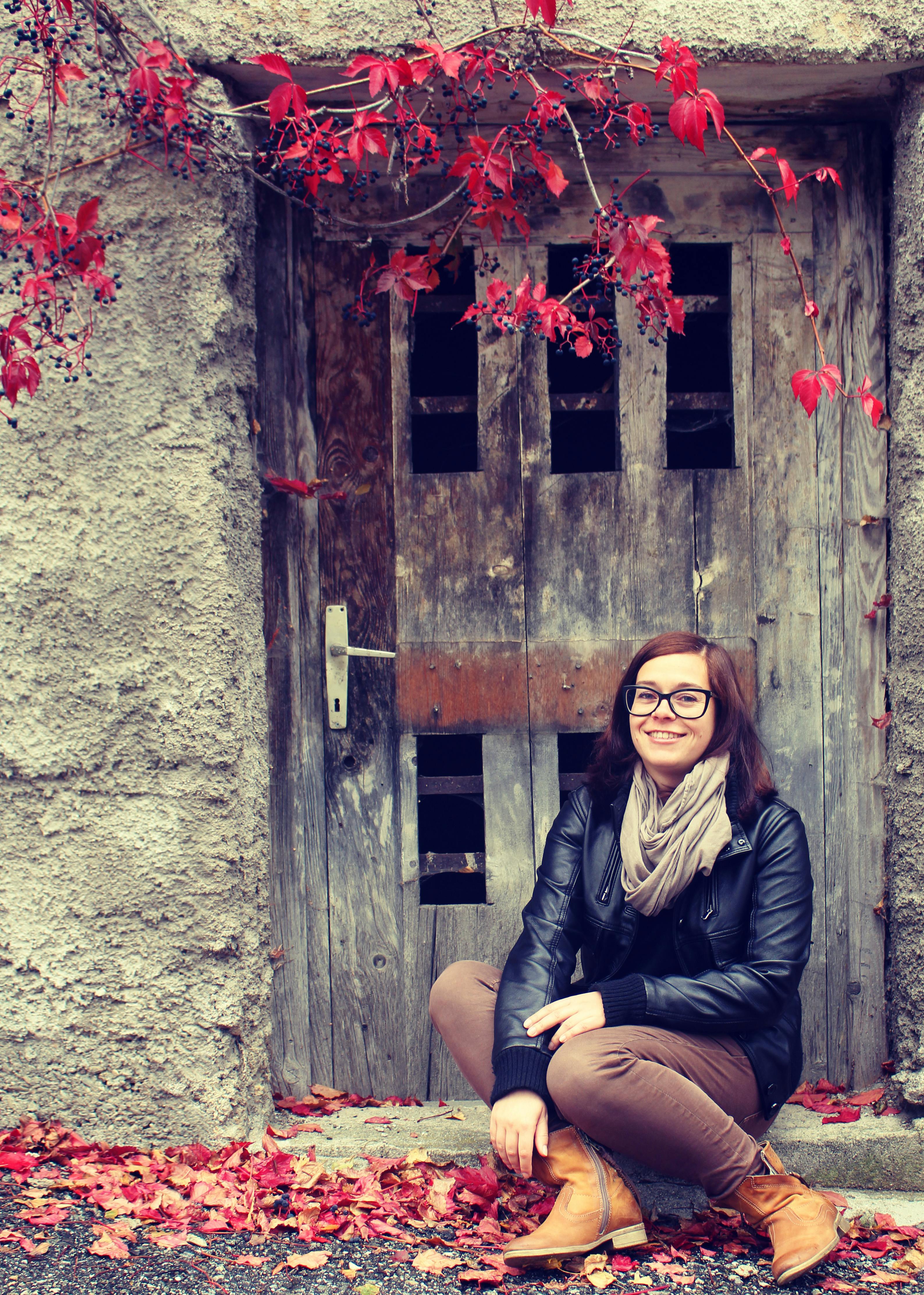 Woman in fall outfit sitting by a rustic door surrounded by red autumn leaves.