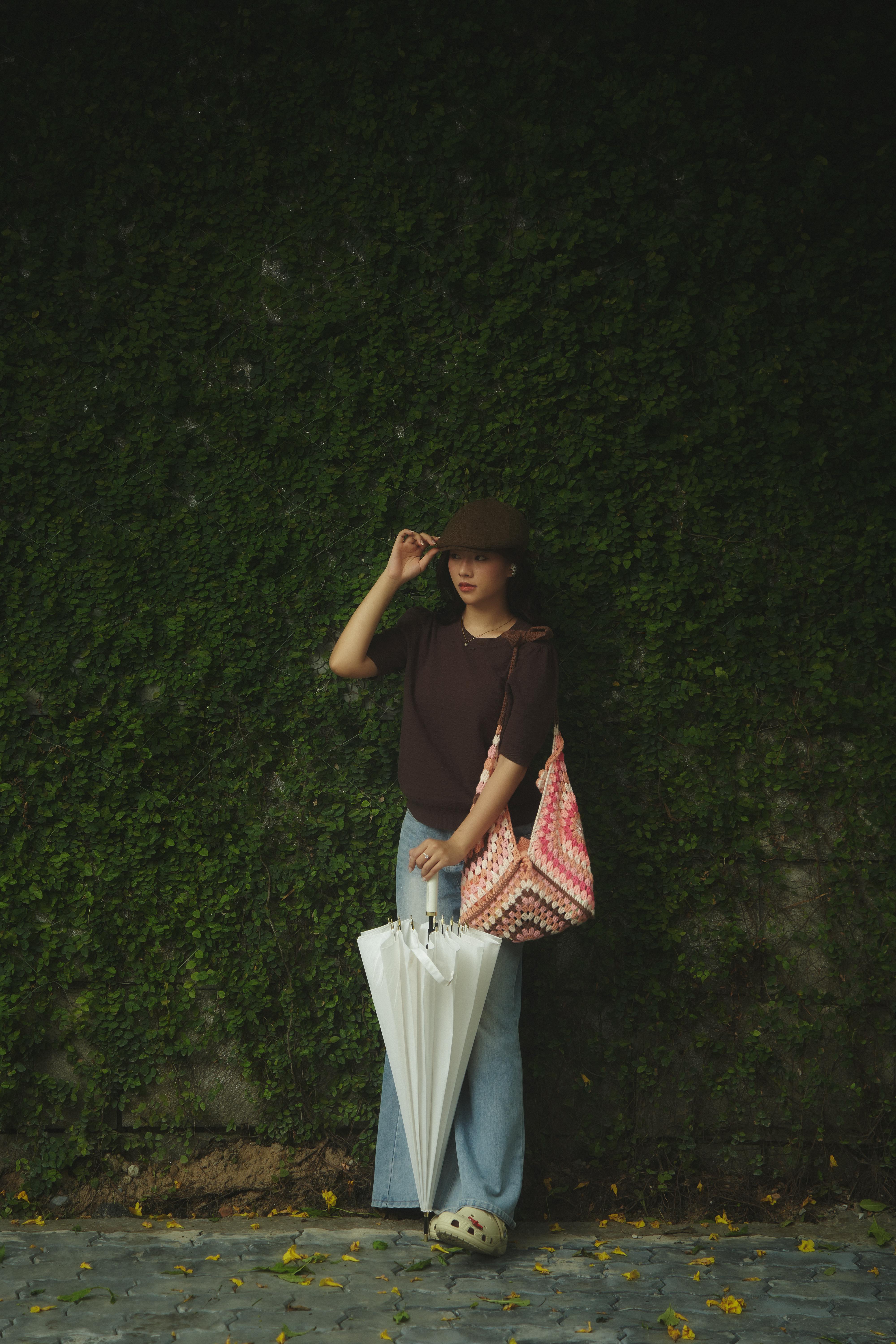 Stylish woman poses by a green wall with an umbrella and hat. Urban fashion vibe.