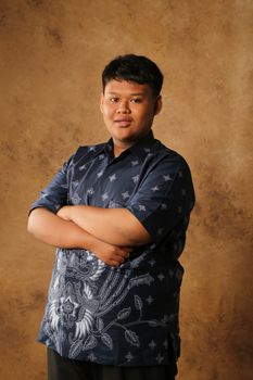 Young man wearing traditional batik shirt poses confidently against a brown textured backdrop. - Fotografer Sidoarjo