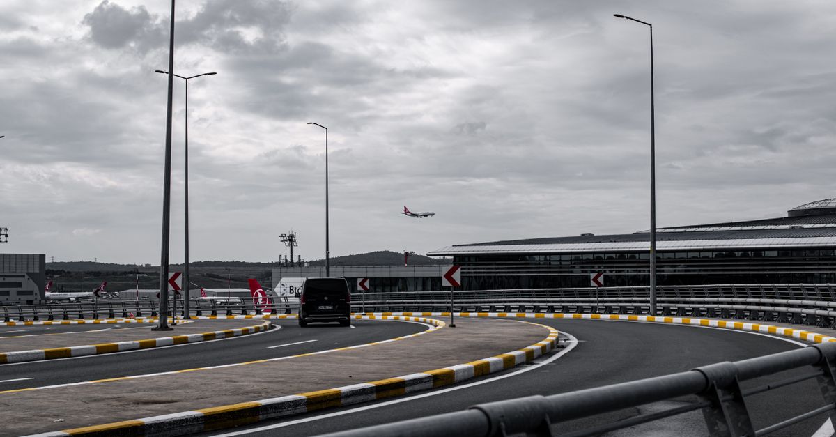 An airplane approaches for landing near a modern airport terminal with a curving road in the foreground.