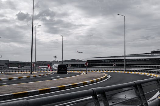 An airplane approaches for landing near a modern airport terminal with a curving road in the foreground.