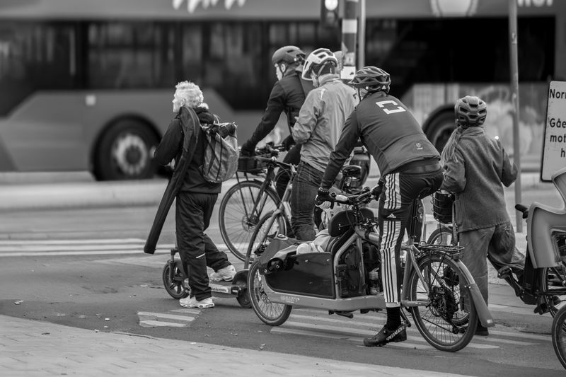 Black and white photo capturing cyclists and scooter riders waiting at a city intersection.