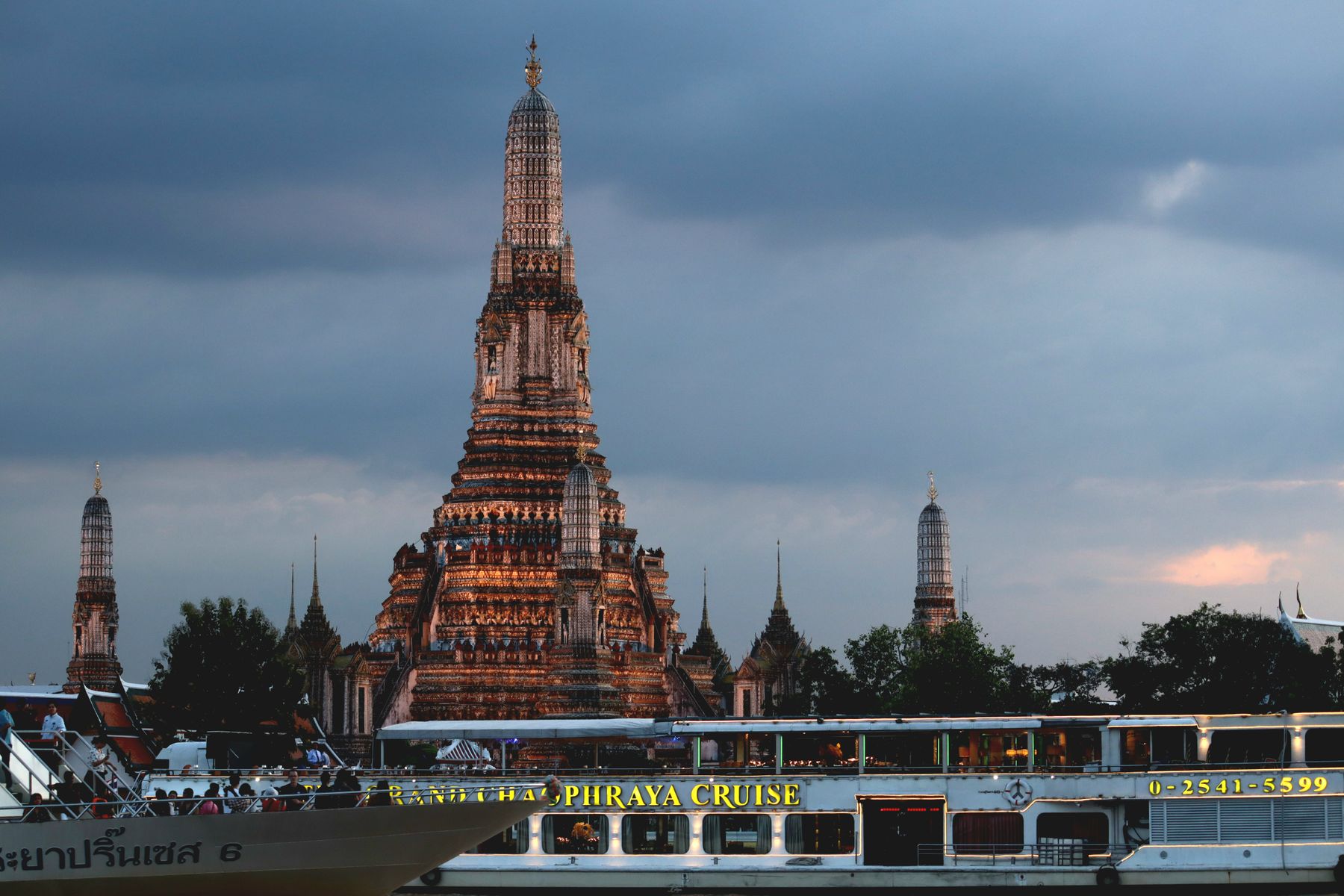 Wat Arun illuminated across the Chao Phraya River at twilight with a passing cruise boat