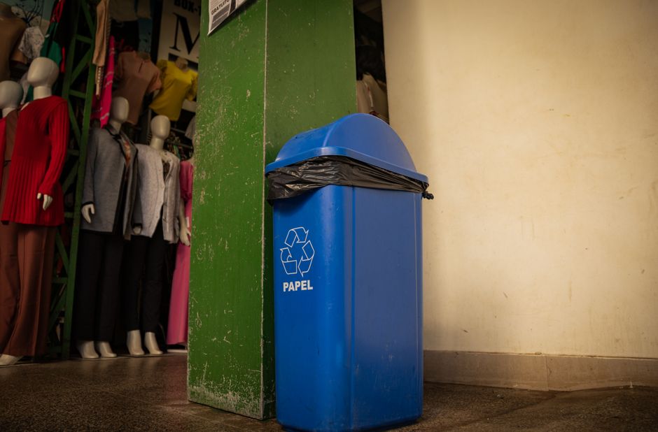 Blue recycling bin labeled 'papel' inside a boutique with mannequins and clothing.