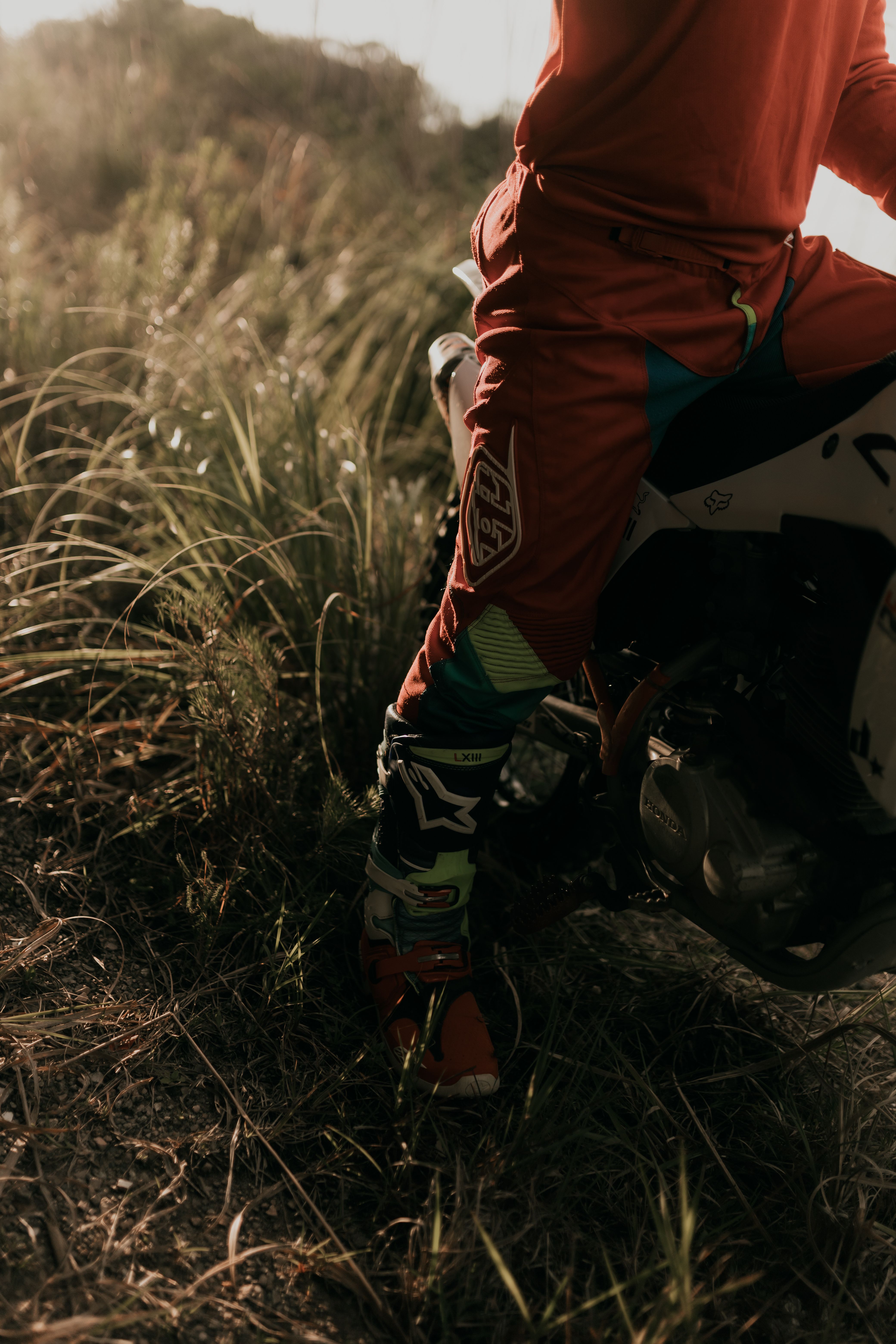 Dirt bike rider in colorful gear with a focus on boots and bike engine in a grassy setting.