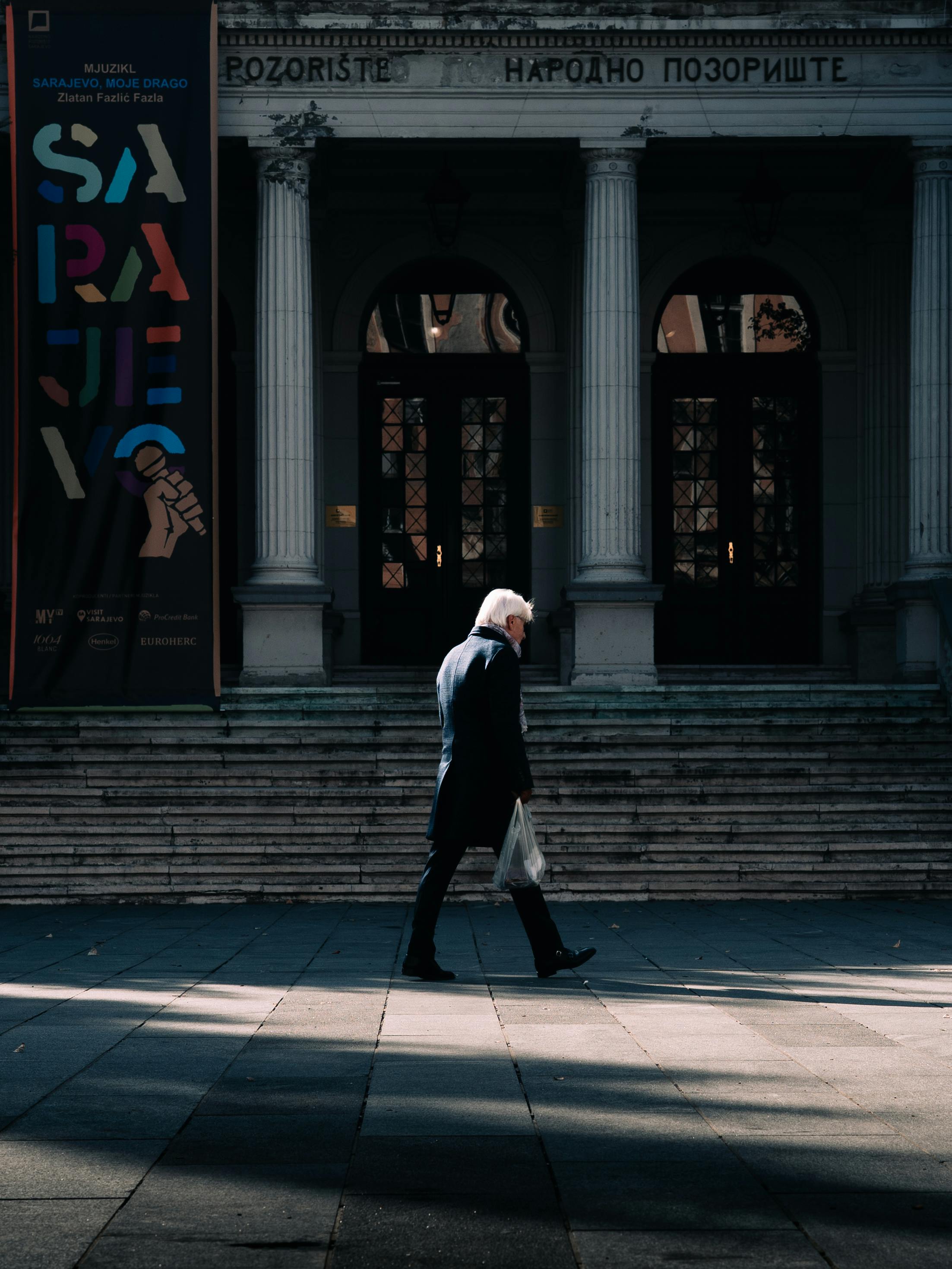 A senior man strolls past the Sarajevo National Theatre, casting a shadow on the pavement.