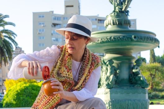 A woman pours mate tea by a historic fountain in Montevideo, Uruguay, enjoying a cultural moment.
