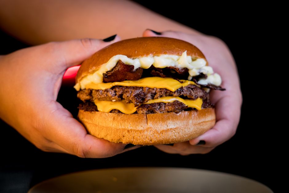 Close-up of a mouthwatering double cheeseburger with bacon, held in hands over a dark background.