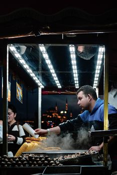 A street vendor tends to roasted corn in a bustling market at night, city backdrop visible.