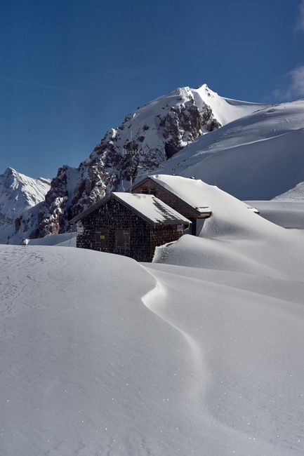 Scenic view of a wooden cabin surrounded by snow-covered peaks in a sunlit alpine setting.
