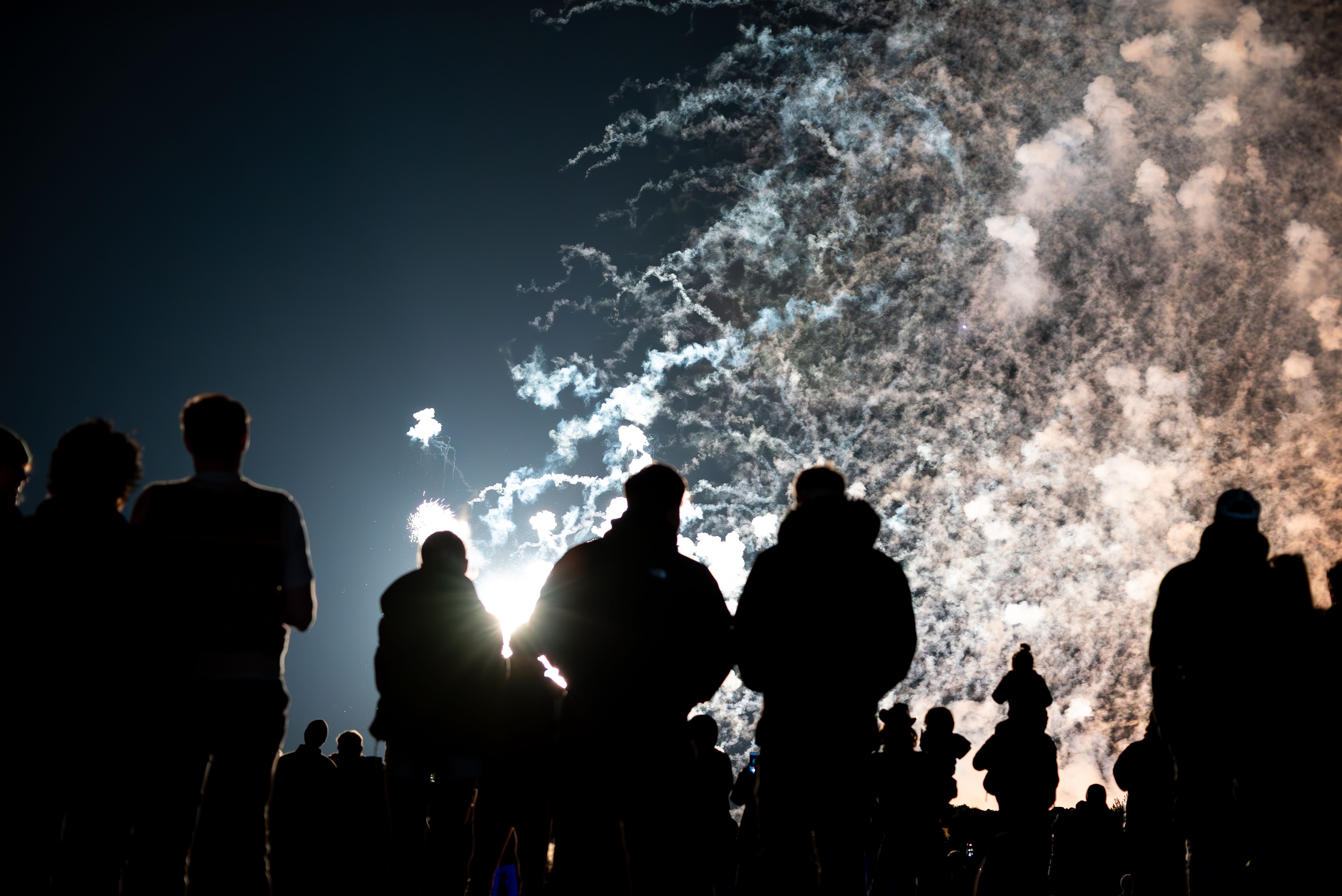 A silhouetted crowd watches fireworks light up a hazy night sky