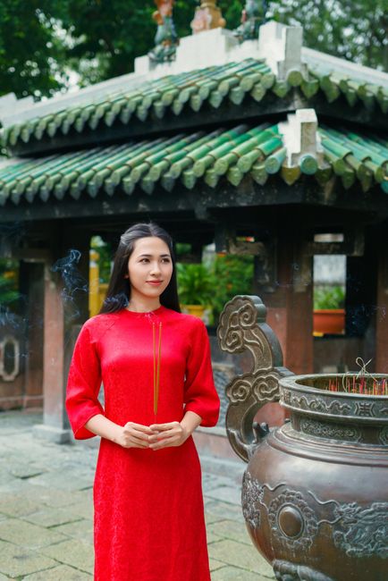 A woman in traditional red áo dài holds incense in front of a historic shrine in Ho Chi Minh City, Vietnam.