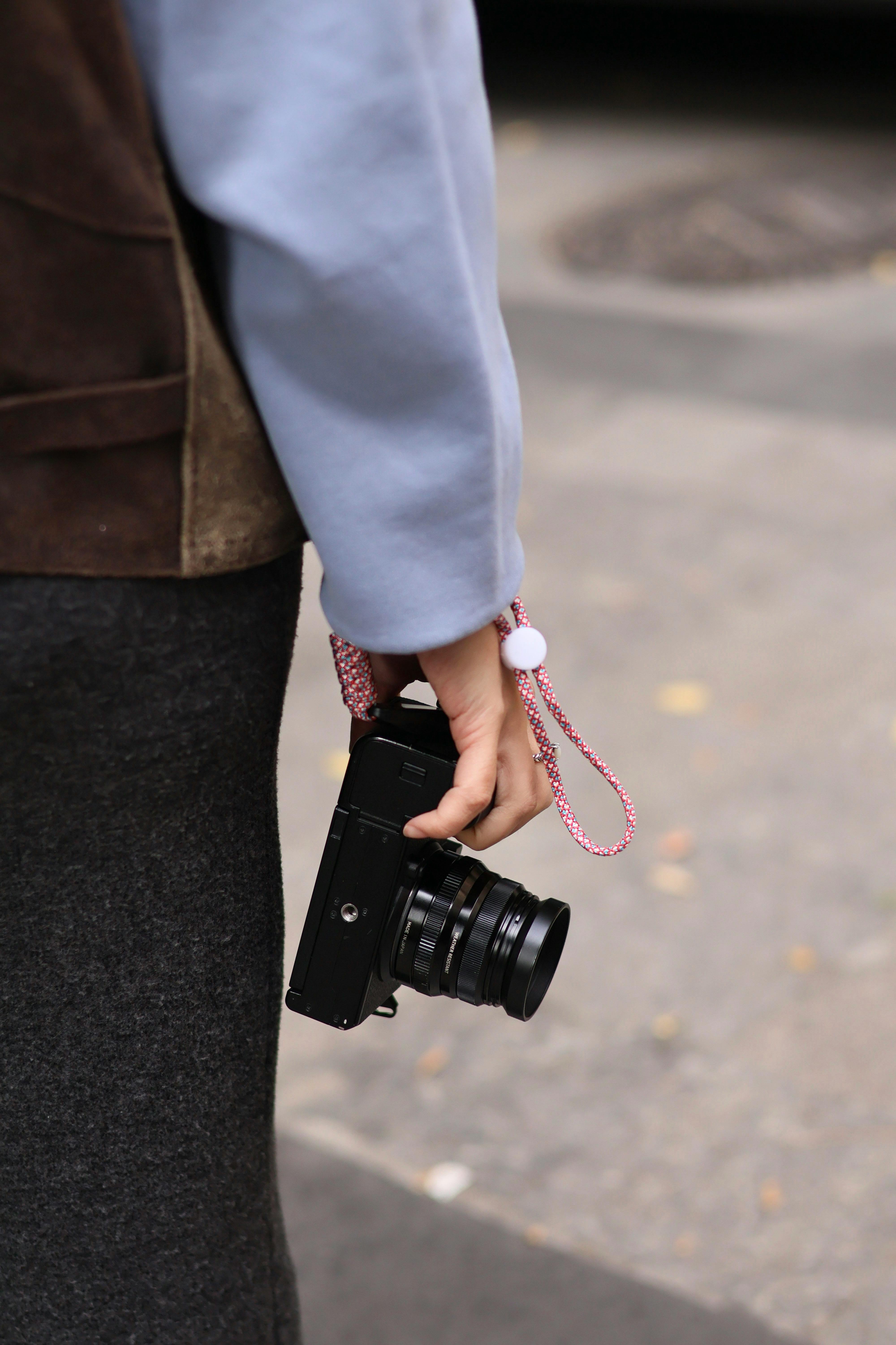 Gratis Una persona che tiene in mano una macchina fotografica nera con un cinturino rosa, immortalata all'aperto. Foto a disposizione