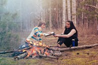 在宁静的森林中，两名妇女围坐在篝火旁，分享着茶与温暖