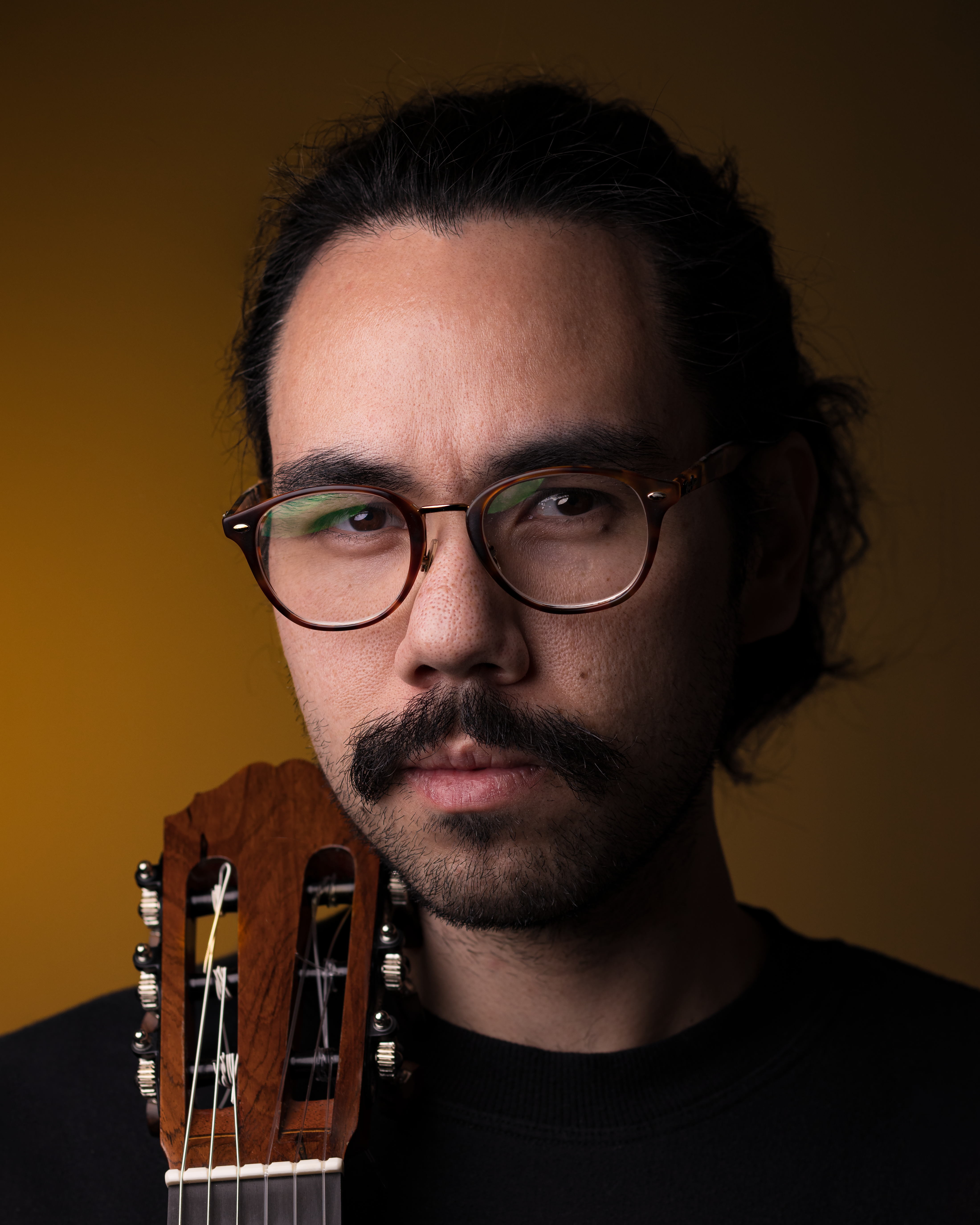 Dramatic portrait of a man with glasses and guitar against a dark background.