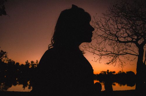 Free A silhouette of a woman by a lake at sunset, surrounded by trees and a vibrant sky. Stock Photo