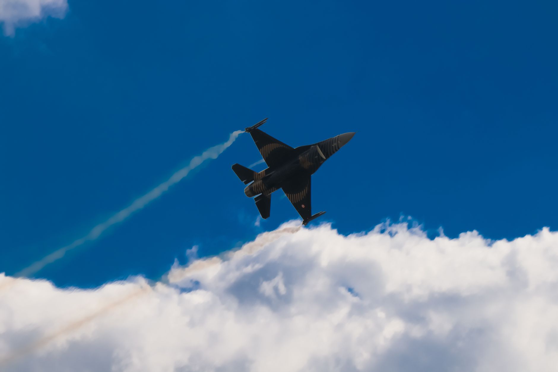 Fighter jet flying in a clear blue sky with clouds below, showcasing speed and power.