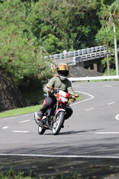 A motorcyclist wearing a helmet rides through a winding road surrounded by lush greenery on a sunny day.