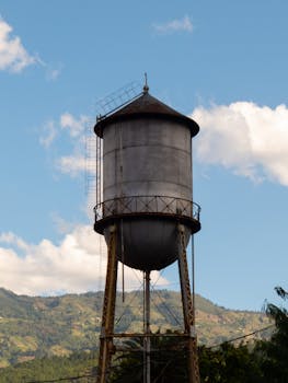 A tall, rustic water tower stands prominently against a scenic mountain background with a blue sky.