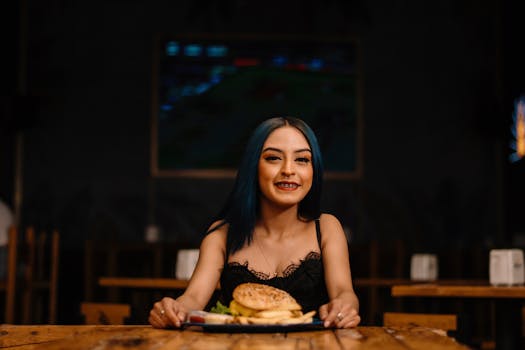 A young woman with blue hair and braces smiles while holding a burger in a cozy restaurant setting.